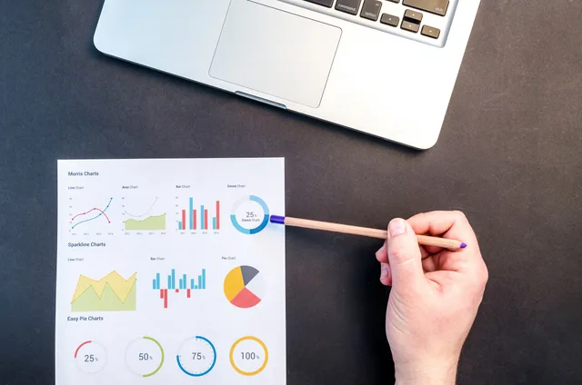 Hand holding pencil reviewing colorful data charts on desk with laptop. Marketing attribution return on investment: a causal method